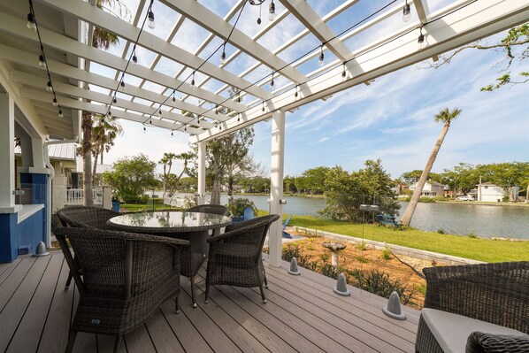 A covered outdoor area with wicker furniture and a glass table, overlooking a lake and palm trees.