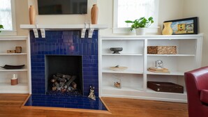 A living room with a blue-tiled fireplace, white shelves, a TV mounted above the fireplace, and a window with a plant and a painting. - Splendor on the Lake, 1928 Cottage-Walk Everywhere (Saint Augustine)