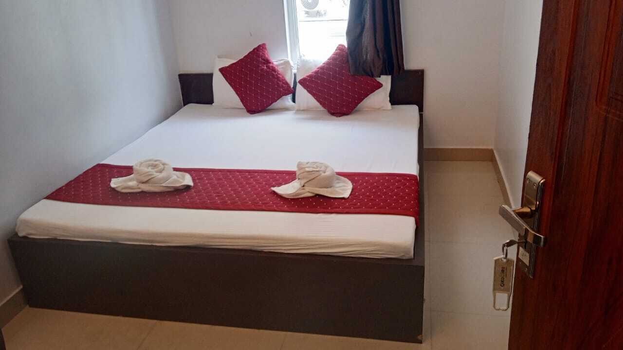 A neatly made bed with a red bedspread and two pillows, a wooden nightstand with a cardholder, and a door with a key lock.