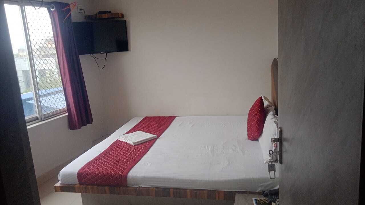 A single bed with a white mattress and red bedding, a small shelf above the bed, and a window with a curtain.