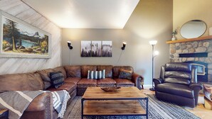 Una sala de estar acogedora con un sofá seccional de cuero marrón, una mesa de centro de madera y un sillón de cuero. Hay luces empotradas en la pared, una chimenea y un cuadro enmarcado con un paisaje.