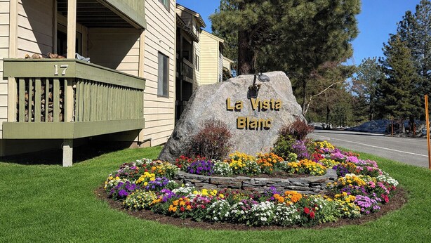 Una gran roca con la inscripción 'La Vista Blanc', rodeada de un macizo de flores de varios colores, frente a un edificio con balcón.
