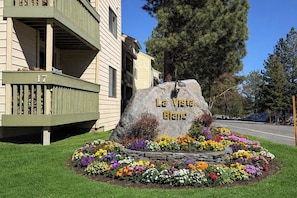 A large rock with 'La Vista Blanc' engraved, surrounded by a flower bed with various colors, in front of a building with a balcony.