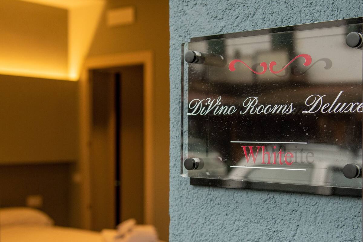 A hotel room with a bed, a wall-mounted sign indicating 'Divino Rooms Deluxe White', and a keypad lock.