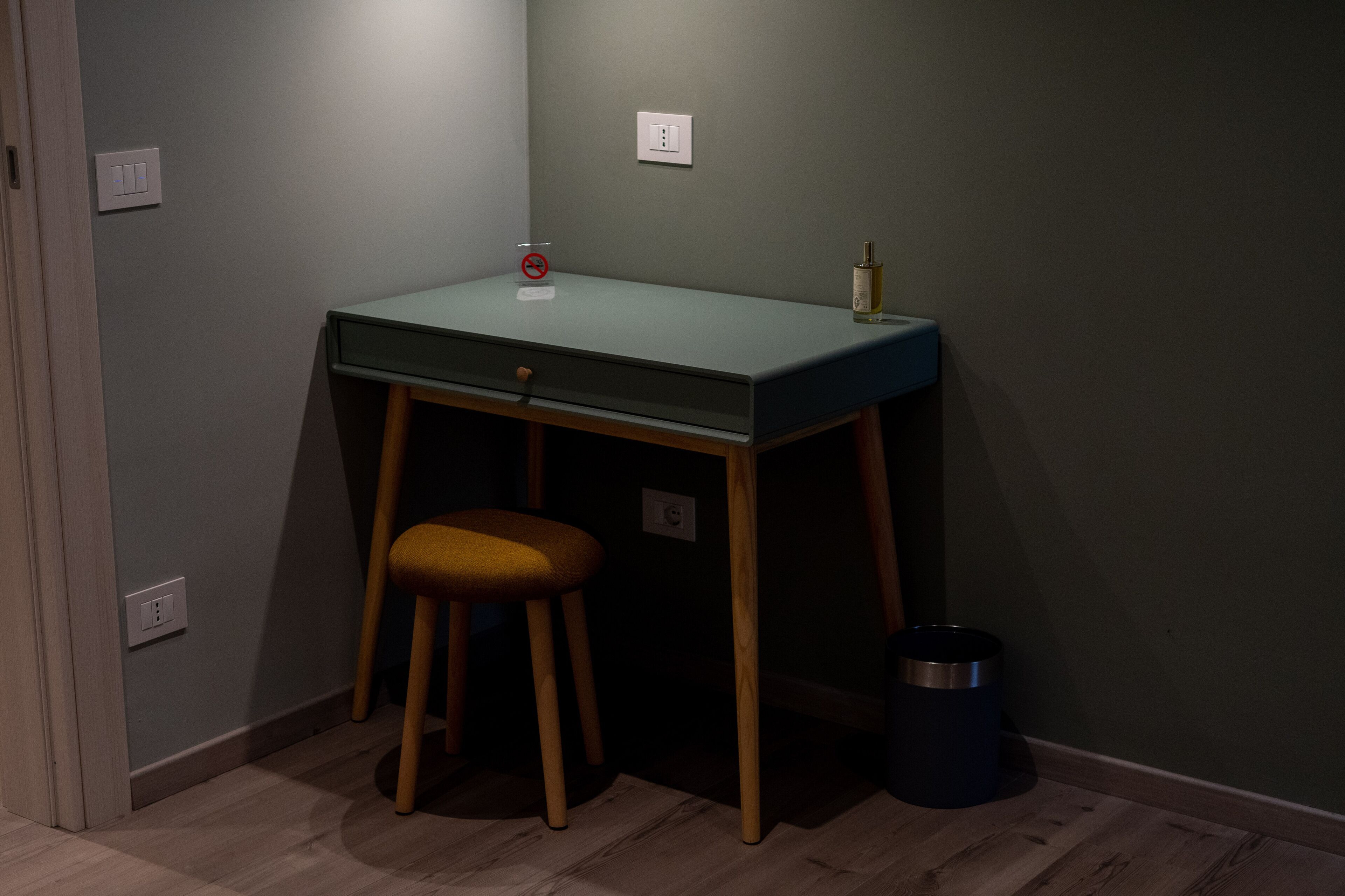 Una habitación pequeña y oscura con un escritorio, un taburete y un bote de basura.