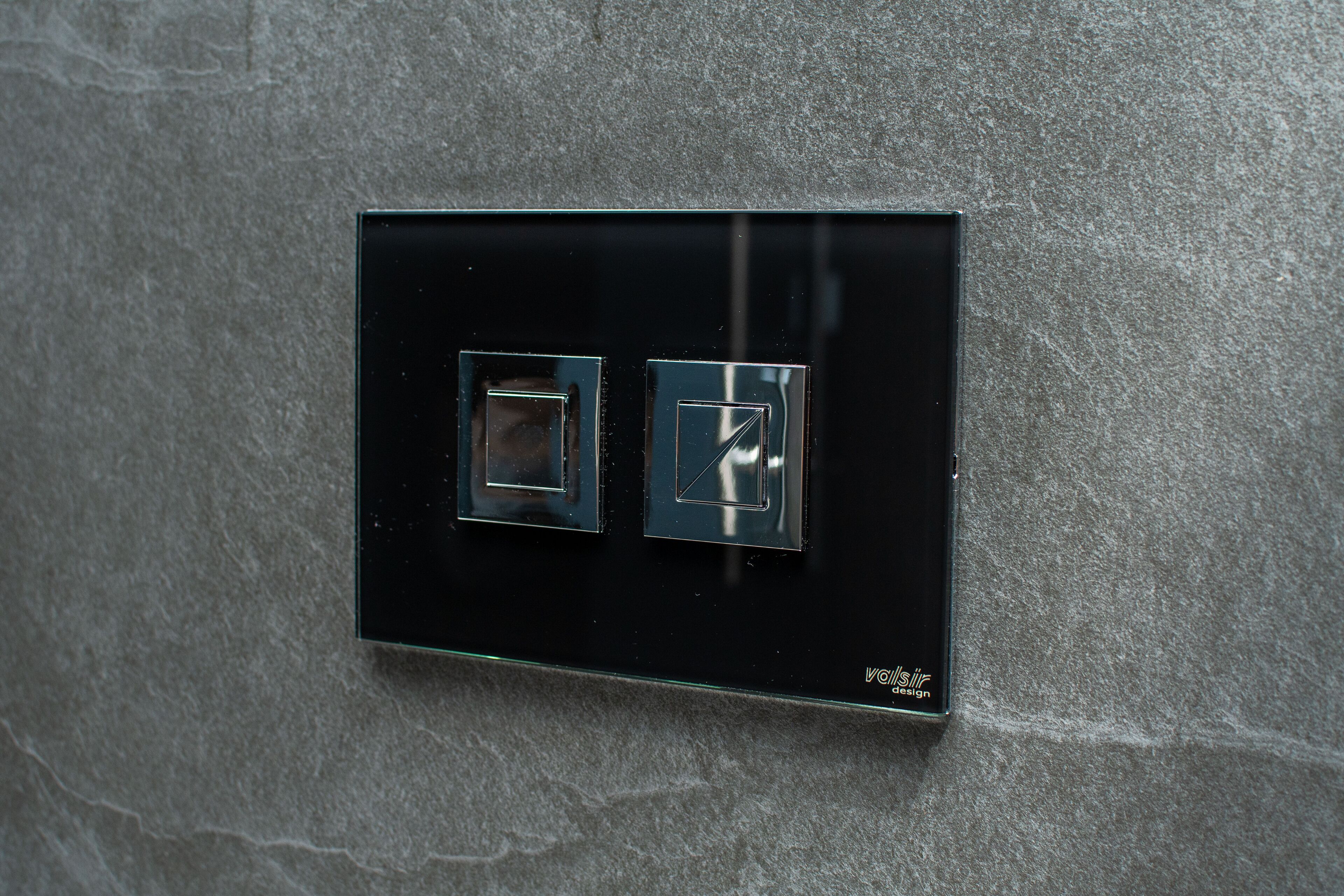 A black switch panel with two square switches on a textured wall.