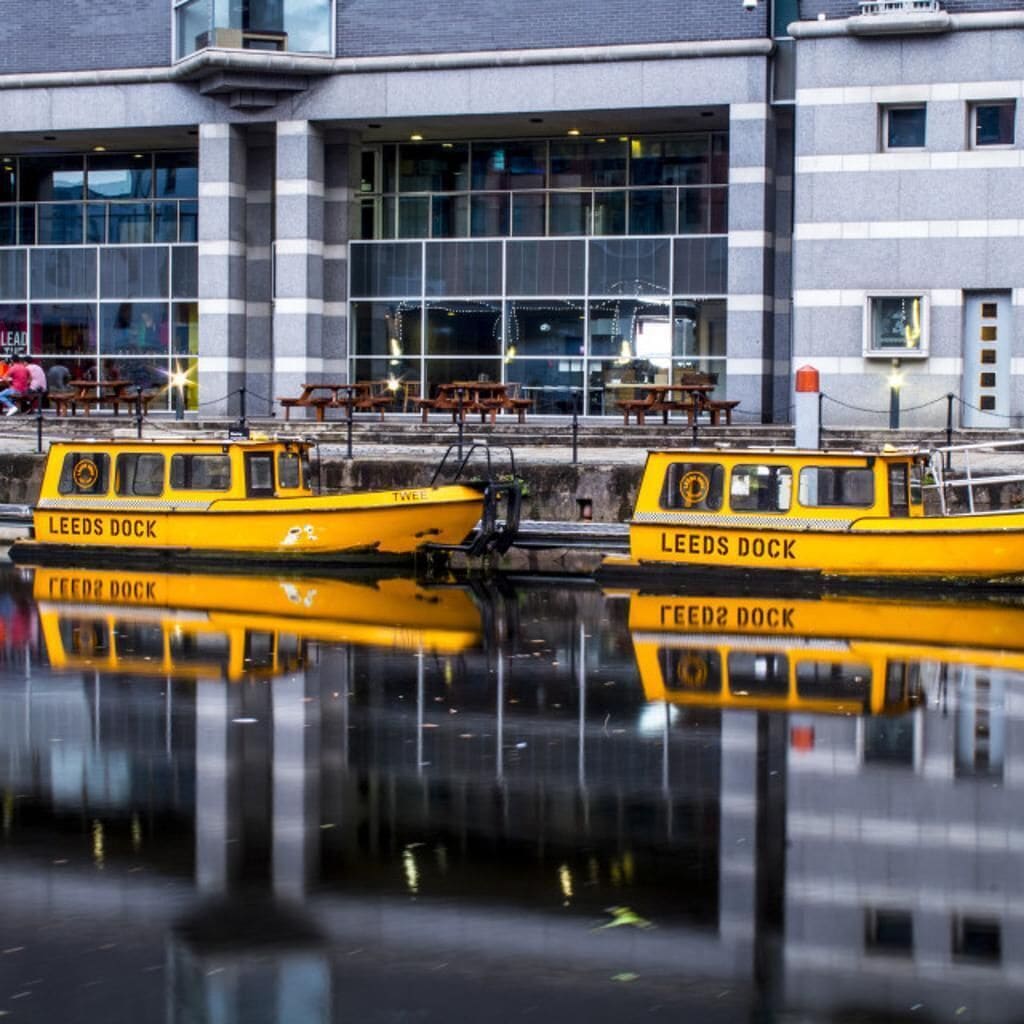 Two yellow boats named 'Leeds Dock' docked at a waterfront with a modern building in the background.