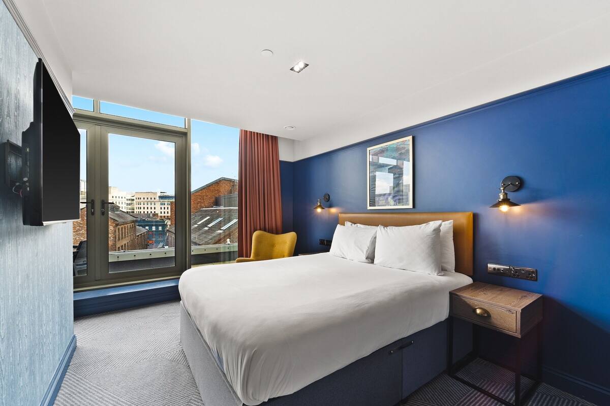 A hotel room with a large bed, a nightstand, a TV mounted on the wall, and a window with a city view.