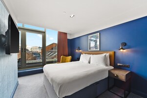 A hotel room with a large bed, a nightstand, a TV mounted on the wall, and a window with a city view.