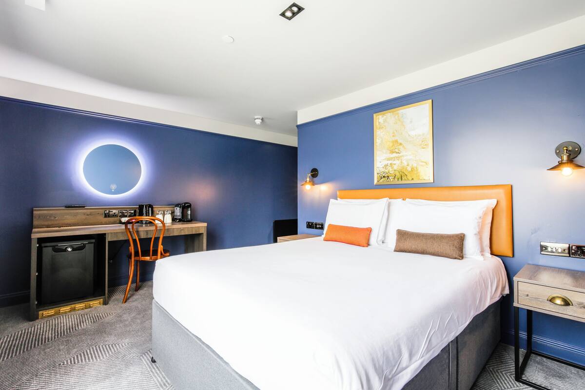 A hotel room with a large bed, a desk with a chair, a small refrigerator, and a wall-mounted light fixture.