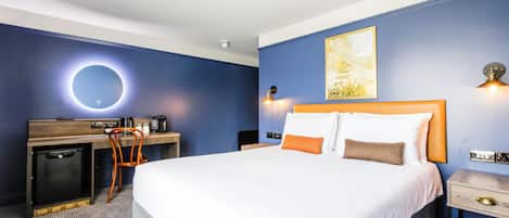 A hotel room with a large bed, a desk with a chair, a small refrigerator, and a wall-mounted light fixture.