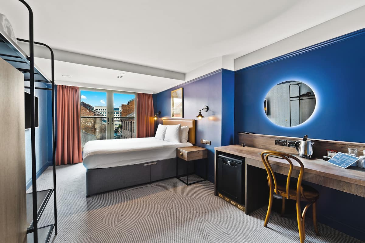 A hotel room with a bed, a desk with a chair, a small kitchen area, and a view of the city through large windows.