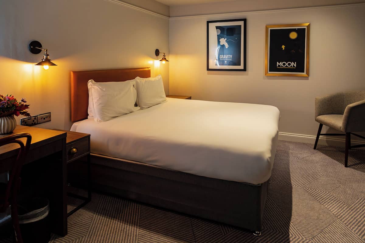 A hotel room with a large bed, bedside lamps, a desk, and a chair. There are two framed pictures on the wall.