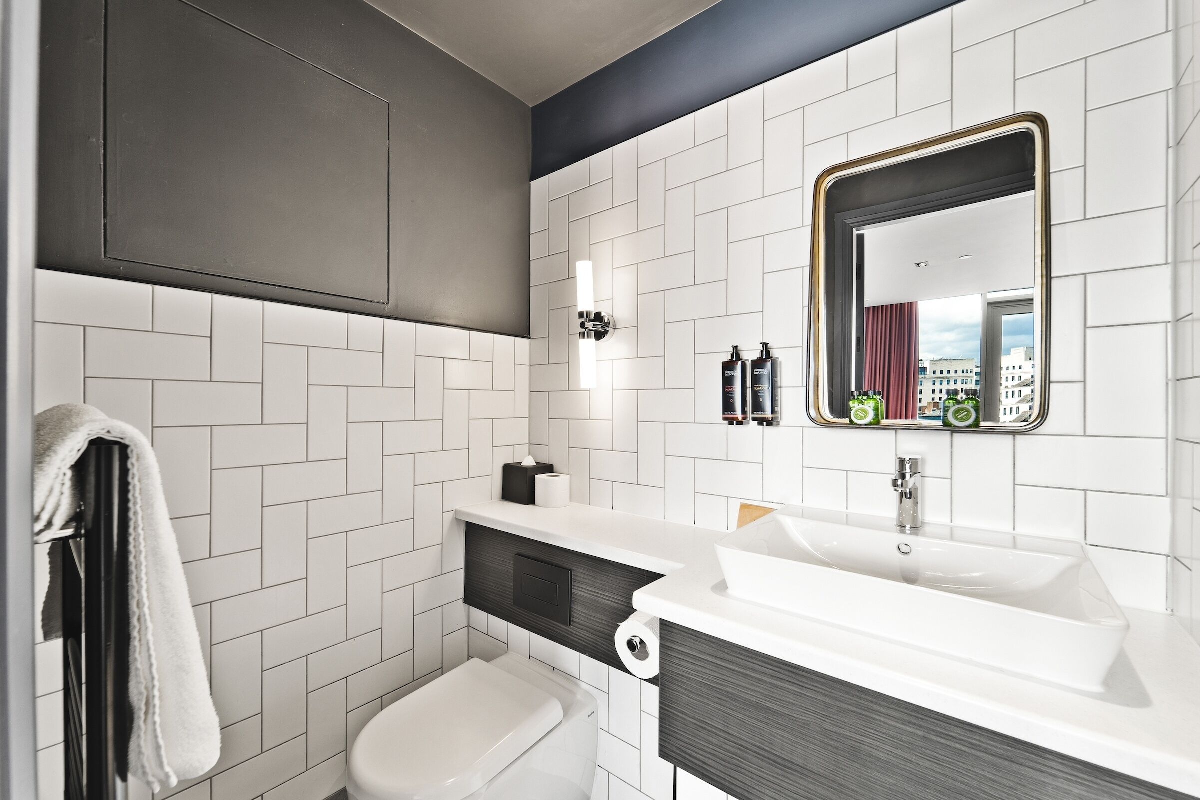 Un baño moderno con baldosas blancas tipo metro, un lavamanos blanco, un mueble de tocador de madera oscura y un espejo grande con marco dorado.