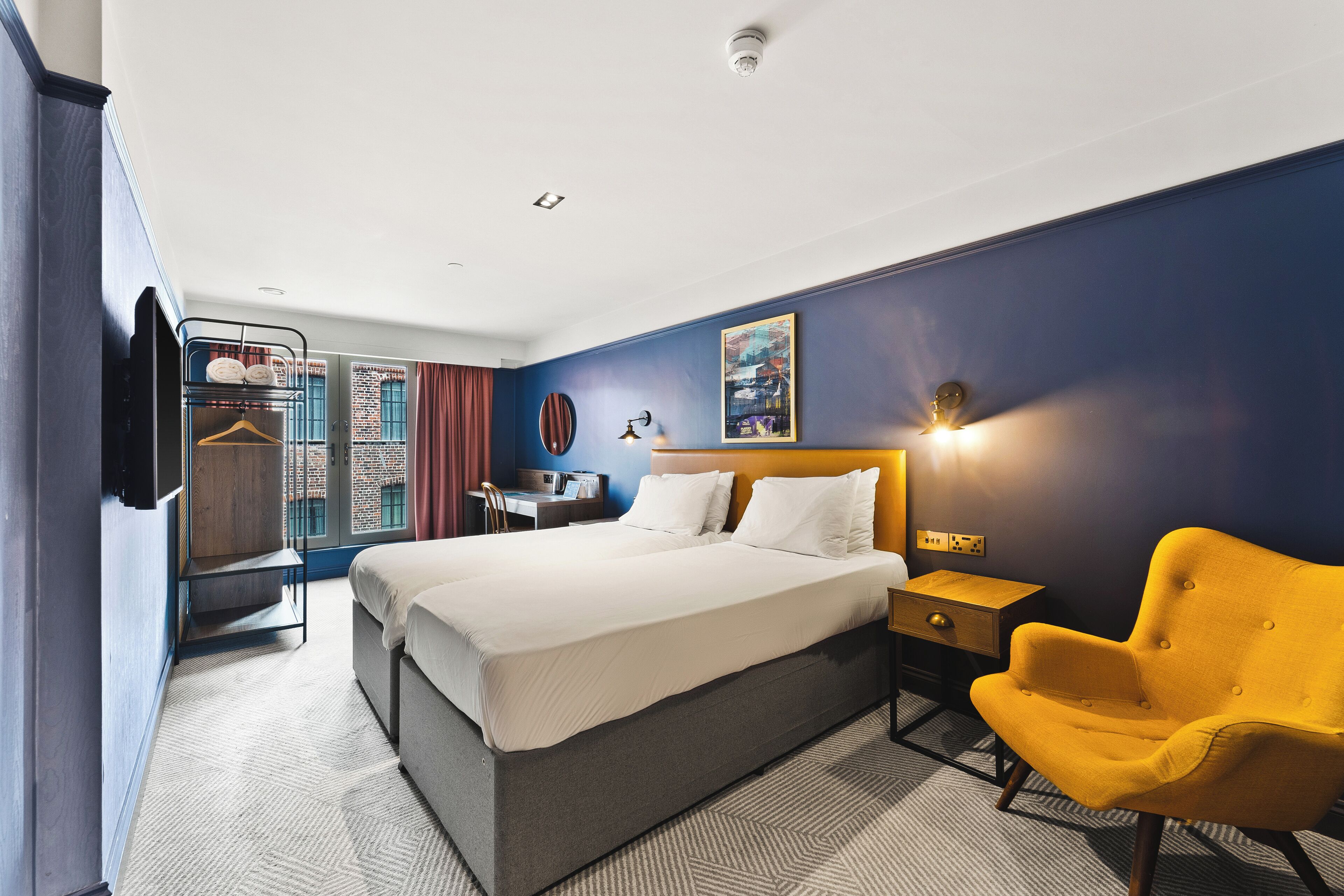 A hotel room with a bed, bedside table, chair, wardrobe, and a painting on the wall.