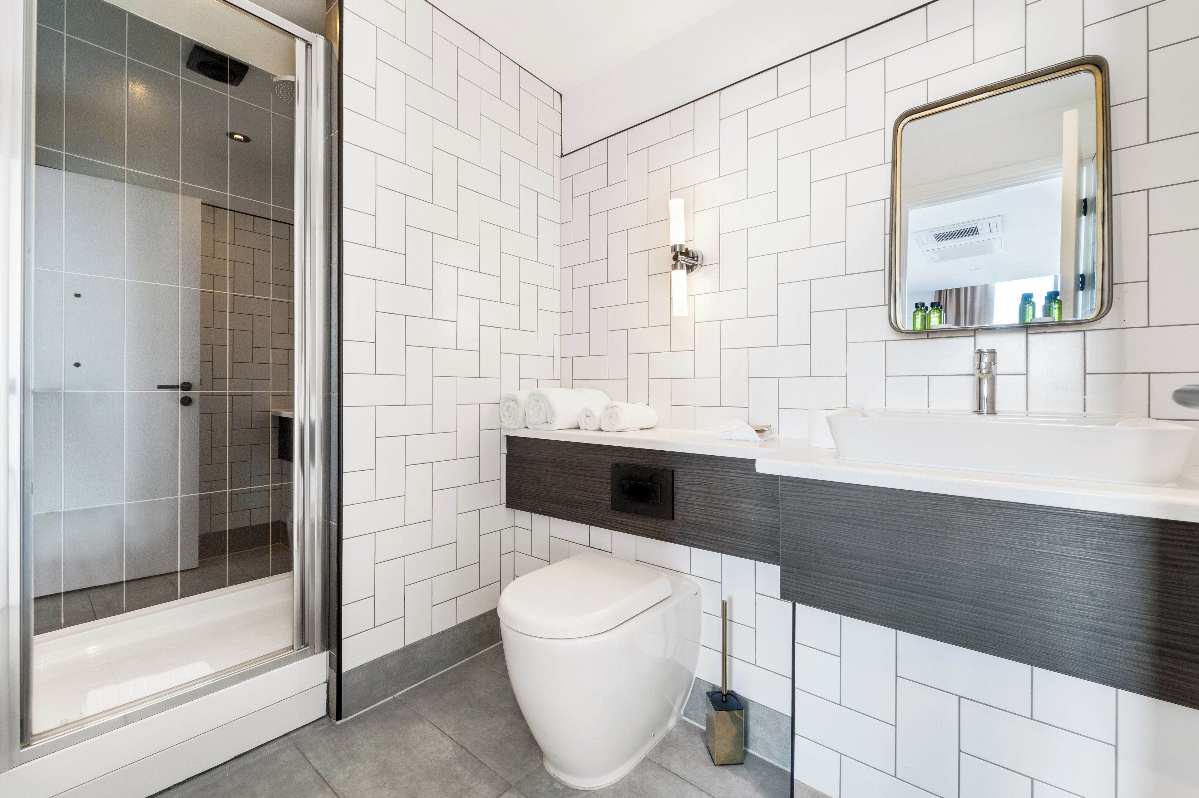 Et moderne badeværelse med hvide flisevægge, et brusekabine i glas, et hvidt toilet, en mørk trækommode og et spejl over vasken.