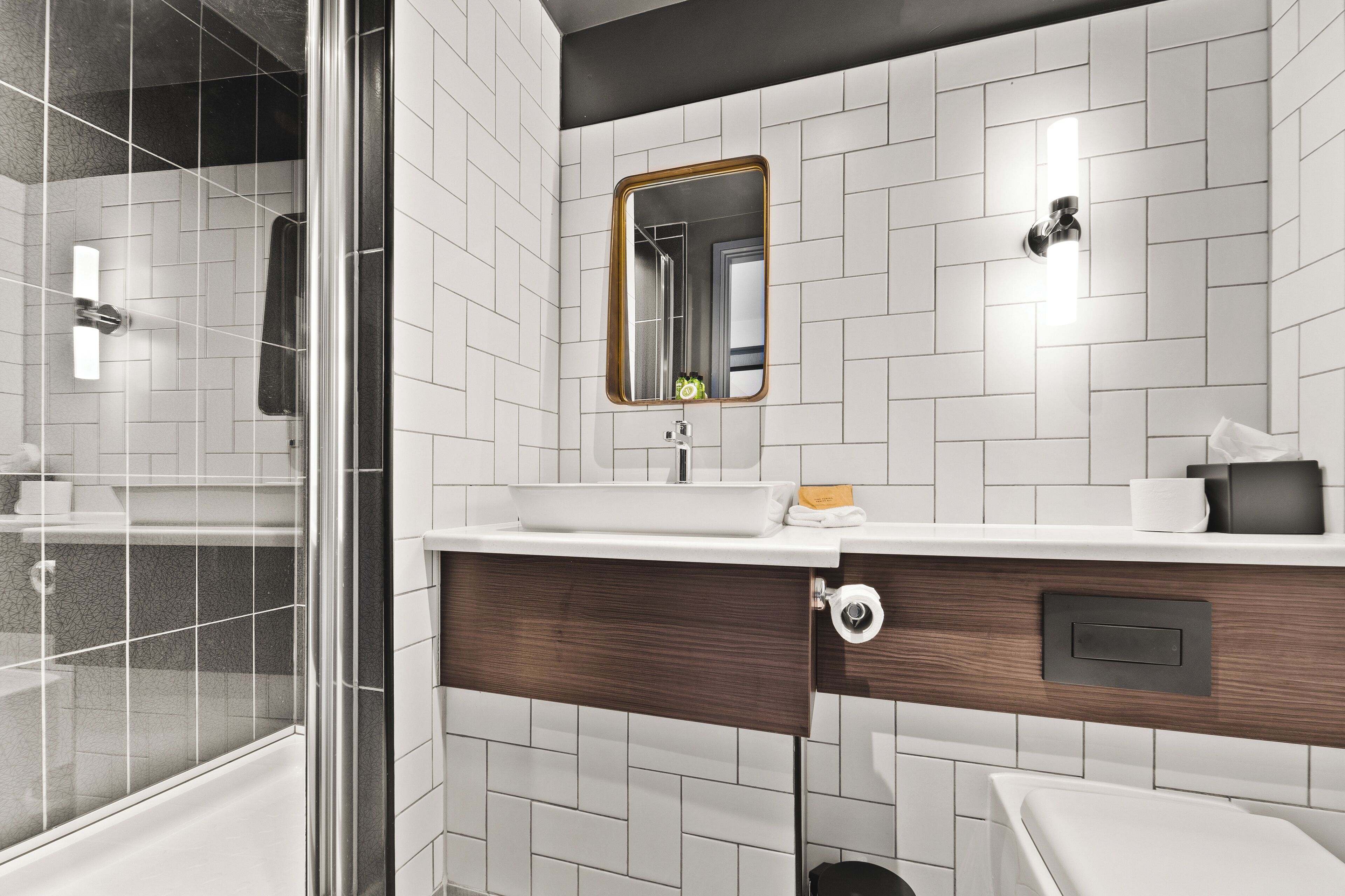 Un baño moderno con paredes de azulejos blancos, un espejo grande, un lavamanos blanco y un mueble de tocador de madera.