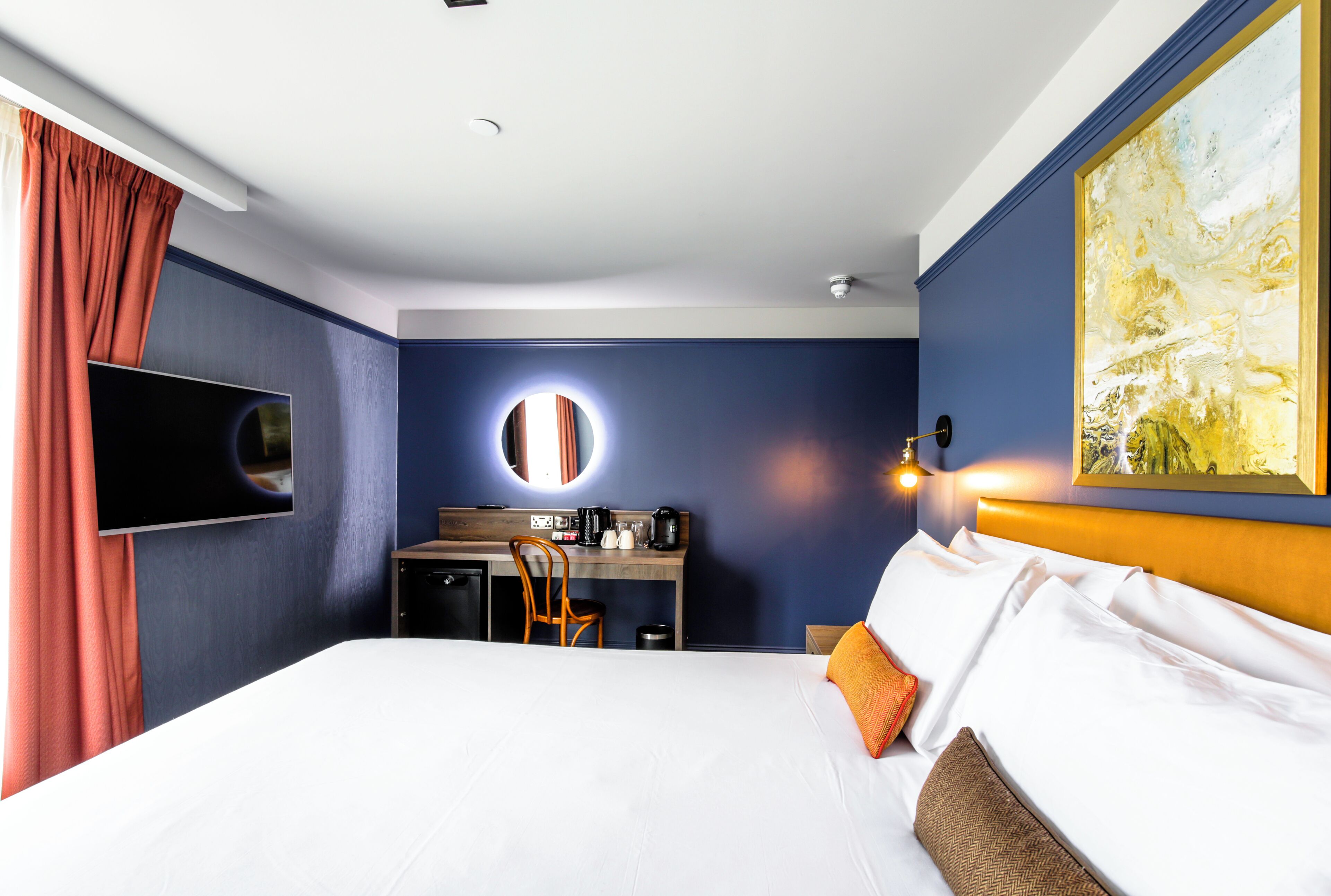 A hotel room with a bed, a desk, a mirror, a television, and a painting on the wall.