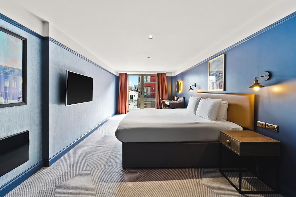 A hotel room with a large bed, a bedside table, a flat-screen TV mounted on the wall, and a window with curtains.
