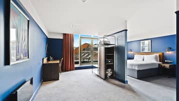 A hotel room with a bed, bedside table, and a desk with a chair. There is a window with a view of buildings outside.