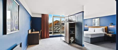 A hotel room with a bed, bedside table, and a desk with a chair. There is a window with a view of buildings outside.