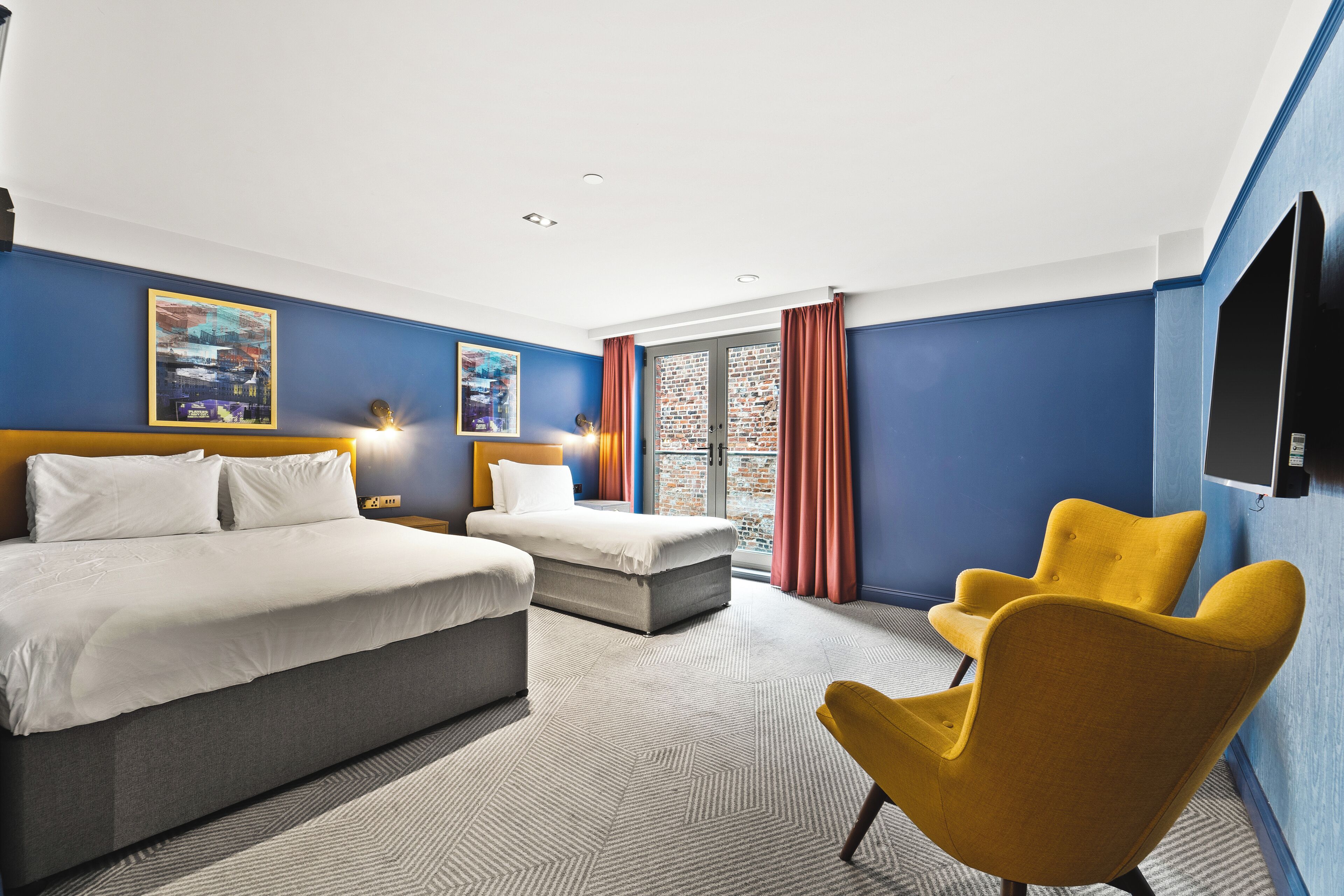 Ein Hotelzimmer mit einem großen Bett, einem kleineren Bett, einem gelben Sessel und Blick auf ein Backsteingebäude durch ein Fenster.