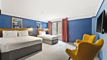 Habitación de hotel con una cama grande, una cama más pequeña, un sillón amarillo y vistas a un edificio de ladrillo a través de una ventana.