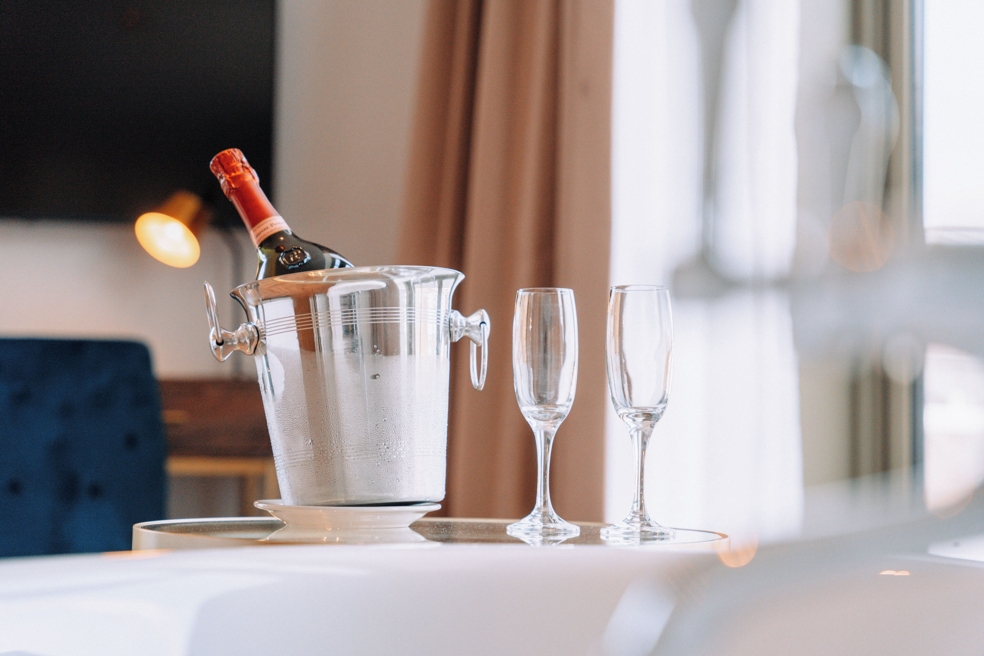A champagne bucket with glasses on a table in a room with a blue chair and a window with curtains.