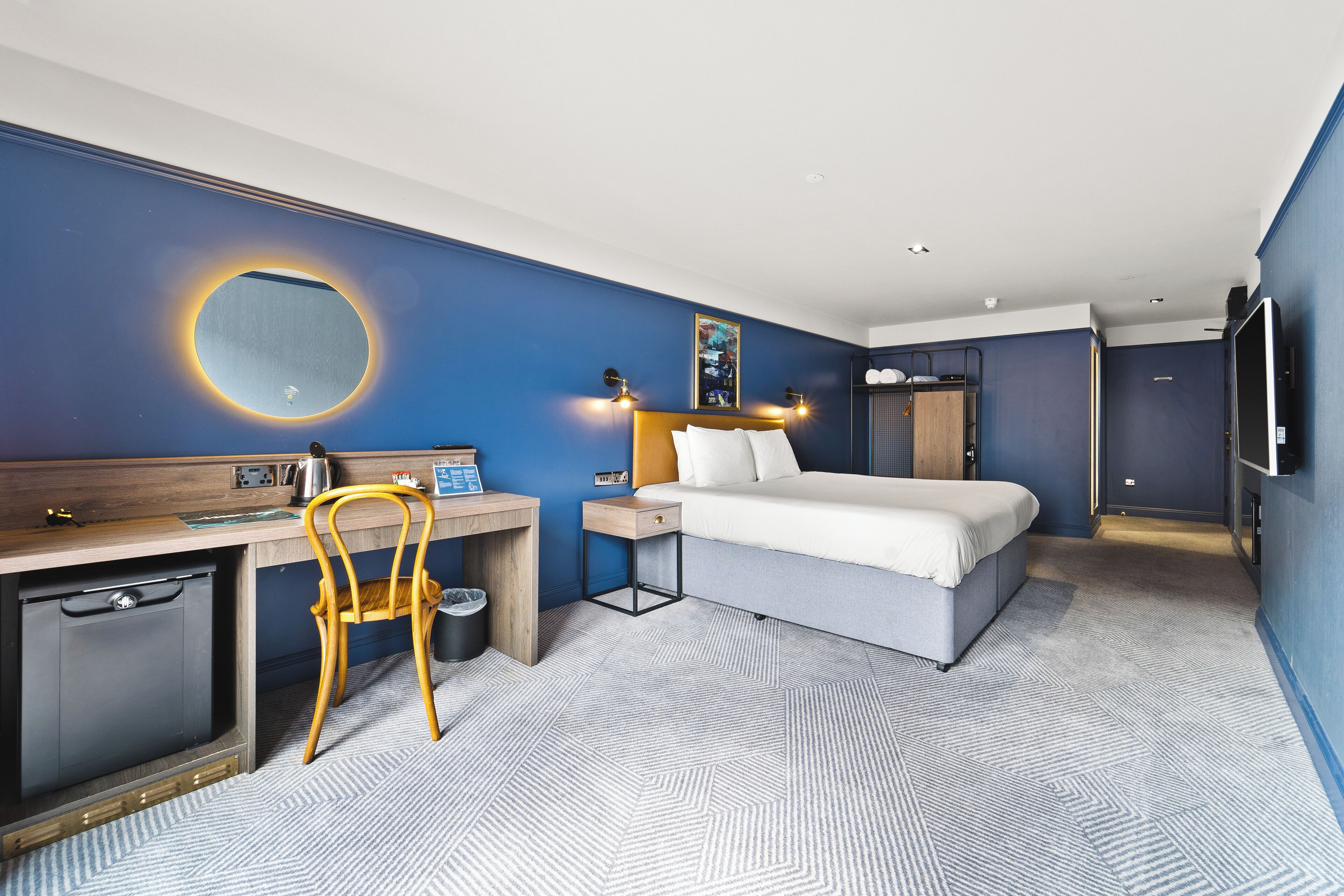 A hotel room with a bed, desk, chair, and a small round mirror.