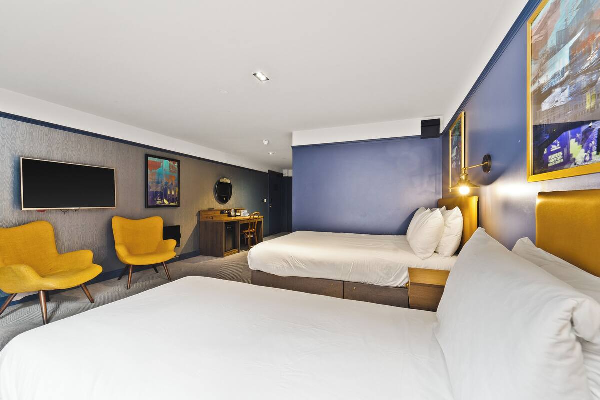 A hotel room with two beds, a desk, two yellow chairs, a flat-screen TV, and artwork on the walls.