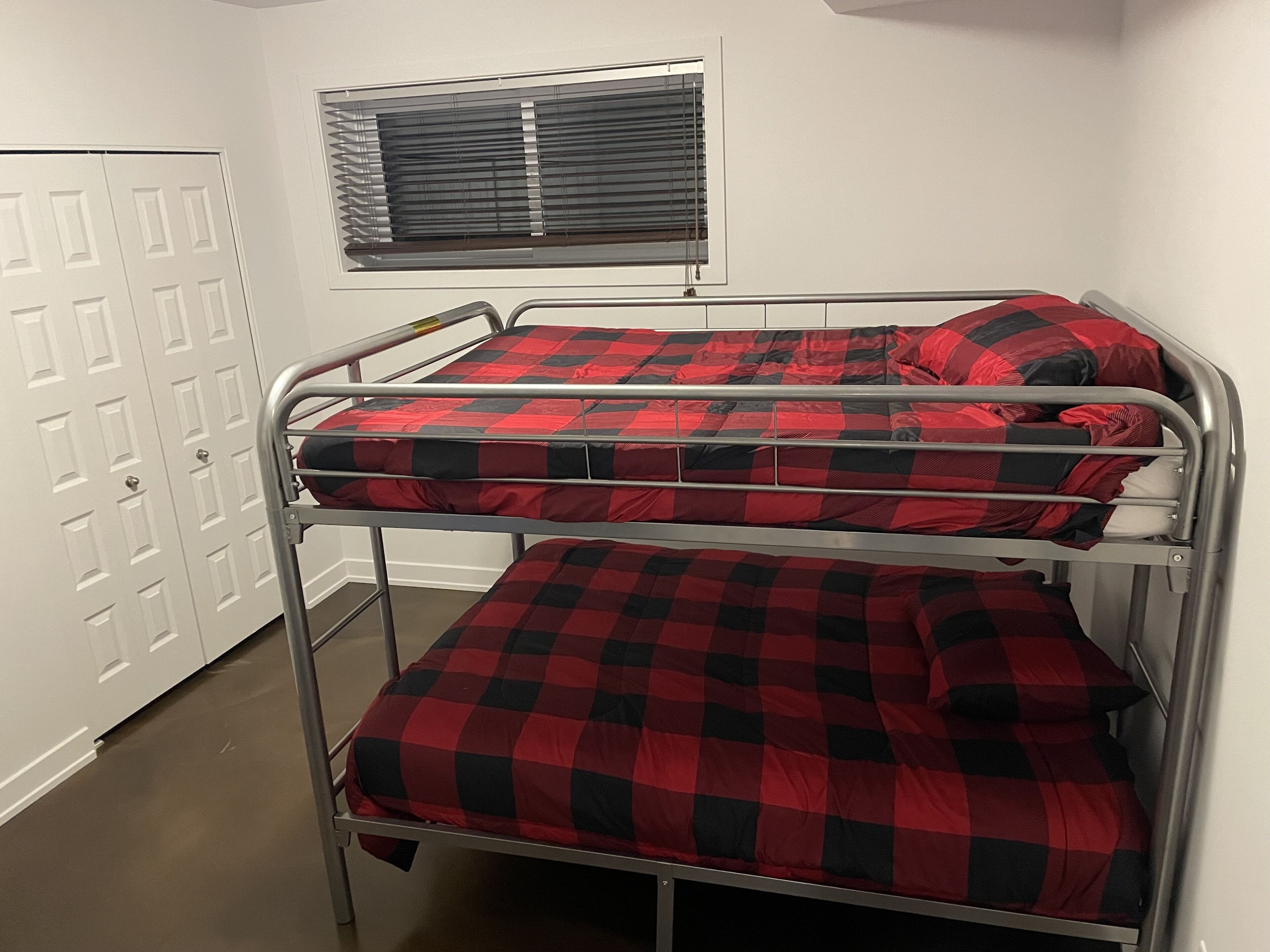 Una litera con ropa de cama a cuadros roja y negra, una ventana con persianas y una puerta blanca.
