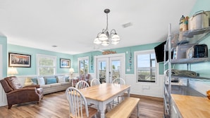 A spacious living area with a wooden dining table, white chairs, a brown leather sofa, and a teal accent wall. - Bayfront Cottage with Screened Porch (Port St Joe)