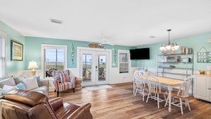 A spacious living room with a brown leather sofa, a striped armchair, and a dining area with white chairs and a wooden table. - Bayfront Cottage with Screened Porch (Port St Joe)