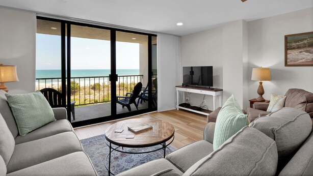 Ein modernes Wohnzimmer mit einer großen Schiebeglasdoor, die auf einen Balkon mit Meerblick führt. Es gibt einen Flachbildfernseher auf einem Standfuß, einen Couchtisch und eine Couch mit Kissen.