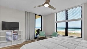 Ein Schlafzimmer mit einem großen Bett, einem Fernseher auf einem weißen Regal, einem Sessel mit Kissen und Blick auf den Strand durch die Fenster.