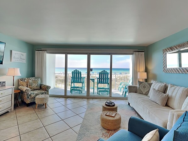 Een woonkamer met uitzicht op het strand, een bank, een fauteuil, een lamp en een kleine tafel.