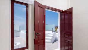 A red door leading to a white-walled room with a view of a pool and outdoor seating. - OUTOPOIA (Santorini)
