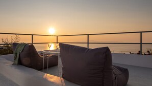 A rooftop terrace with two cushioned seating areas, a table with two glasses, and a view of the sunset over the sea. - OUTOPOIA (Santorini)
