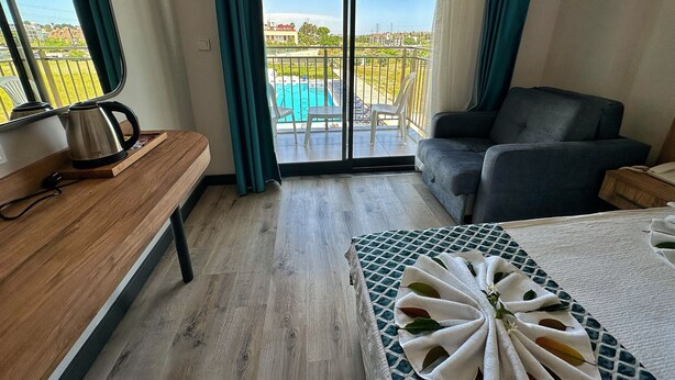 Quarto de hotel com um banco de madeira, uma mesa com uma toalha, um sofá e vista para uma piscina através de uma porta de correr.