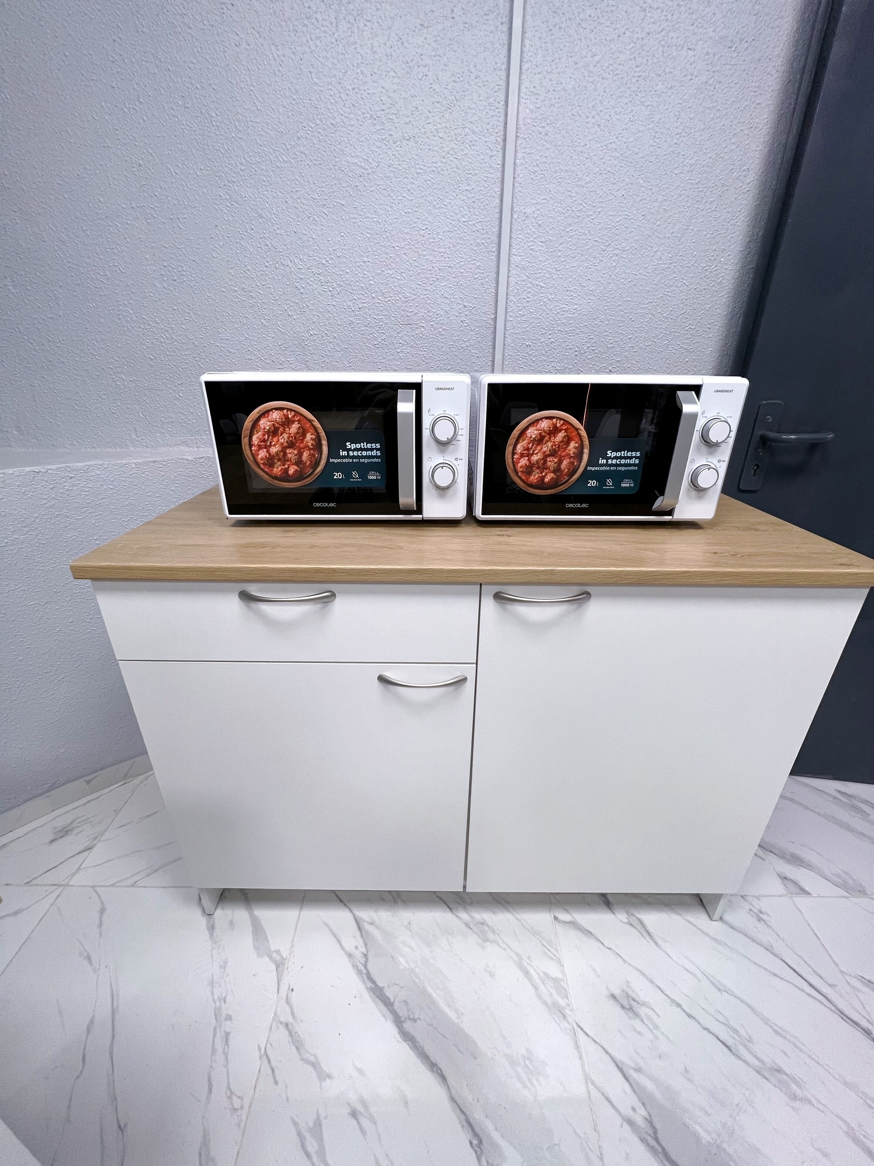 Two wall-mounted microwaves on a wooden countertop above a white cabinet with drawers.