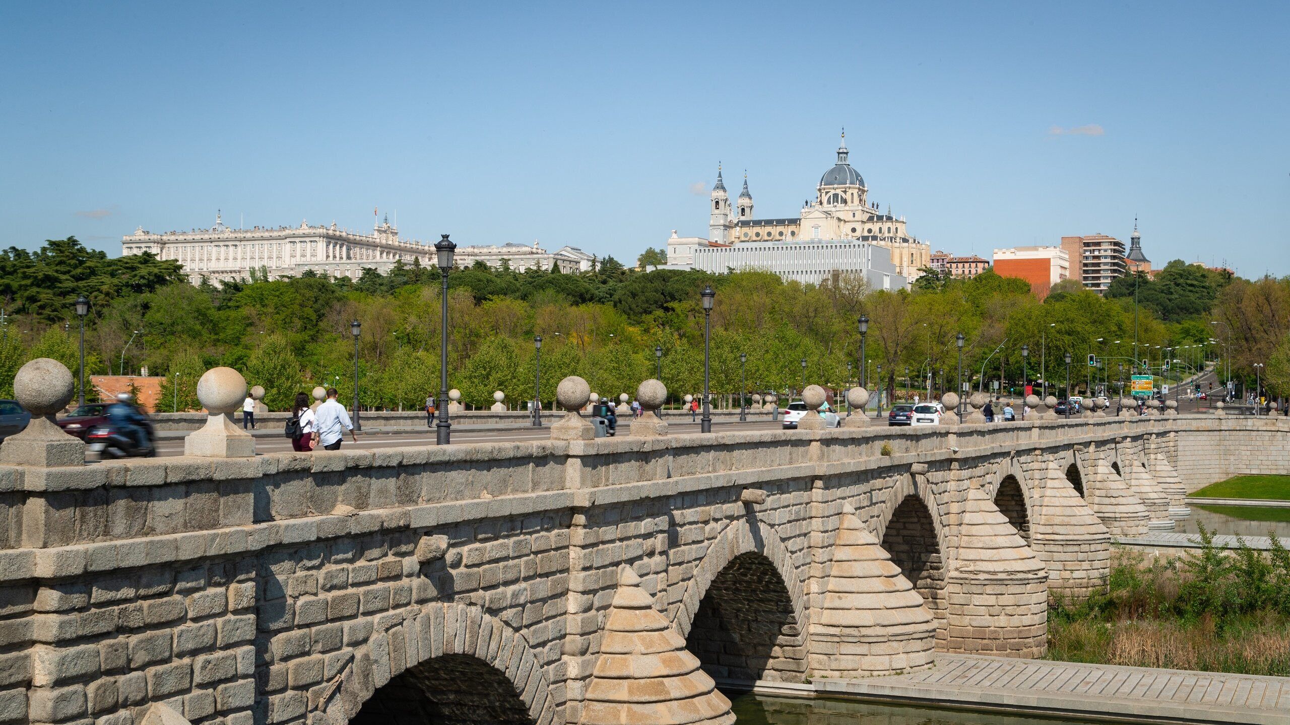 Un puente de piedra con arcos, un río y un paisaje urbano con edificios históricos al fondo.