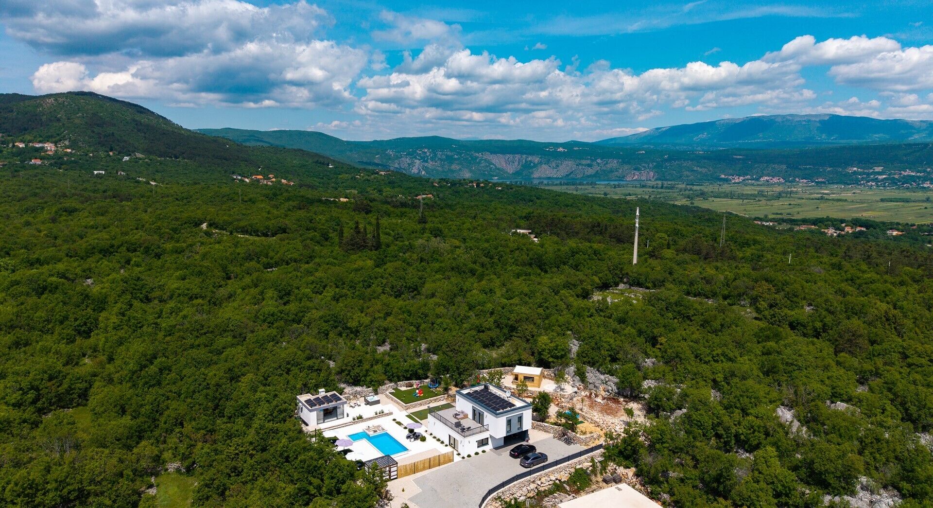 A modern house with a swimming pool, surrounded by dense forest and mountains.