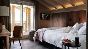 A hotel room with a large bed, a desk with a chair, a television, and a view through a sliding door. - Chalet Le Prariand (Megève)