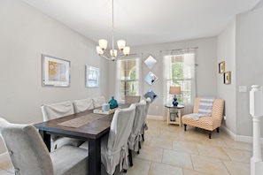 A dining area with a wooden table, grey upholstered chairs, and a chandelier. There are framed pictures on the walls and a small side table with a lamp.