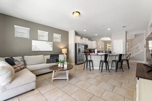 A modern living room with a sofa, coffee table, and a kitchen area with a refrigerator, microwave, and bar stools.
