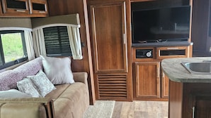 Un intérieur de camping-car compact comprenant un coin cuisine, un espace salon avec un canapé et des coussins, et un grand téléviseur.