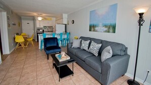 Una sala de estar con un sofá gris, una mesita de centro y sillas amarillas y azules. Se observa una cocina al fondo con armarios blancos y una encimera azul.