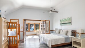 A spacious bedroom with a large bed, a desk, and a chair, a ceiling fan, and a view of the outdoors through large windows. - Seacluded Tree House, Oceanfront House in Pine Knoll Shores, NC! (Pine Knoll Shores)