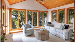 A spacious living room with a high ceiling, large windows, a white sofa, a chair, and a woven ottoman. - Seacluded Tree House, Oceanfront House in Pine Knoll Shores, NC! (Pine Knoll Shores)
