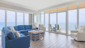 Uma sala de estar ampla com um sofá azul grande, uma mesa de centro e vista para o oceano através de grandes janelas.